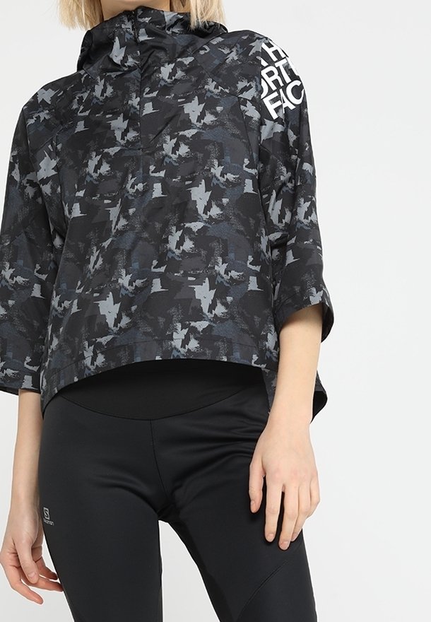 Femme portant un pull à motif camouflage noir et gris à manches trois-quarts et un legging de sport noir sur fond blanc.