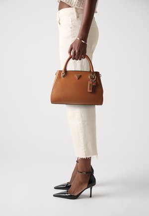 Sac à main en cuir tan avec un design structuré, dotée d'une poignée supérieure, de quincaillerie en argent et d'une étiquette de marque. Porté avec un pantalon blanc et des talons noirs.