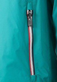 Petrolgroene jas met een tweekleurige ritssluiting; de buitenste laag is glad met een ruitpatroon, en de ritssluitingaccenten zijn rood en wit.