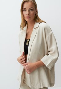 Beige Leinenblazer mit entspannter Passform, mit Reverskragen und Dreiviertelärmeln. Kombiniert mit einem schwarzen Oberteil und goldenen Schmuck.