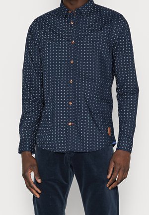 Mężczyzna ubrany w granatową koszulę z długim rękawem, ozdobioną małymi białymi wzorami geometrycznymi i brązowymi guzikami, w połączeniu z ciemnoniebieskimi spodniami.
