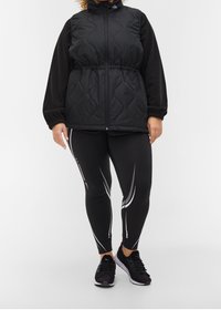 Schwarze gesteppte Weste mit hohem Kragen, tailliertem Schnitt und langen schwarzen Ärmeln. Schwarze Leggings mit weißen Akzentstreifen. Schwarze Sportschuhe.