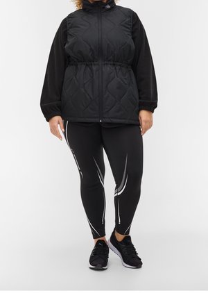 Gilet noir matelassé avec col montant, taille cintrée et longues manches noires. Leggings noirs avec des bandes accentuées blanches. Chaussures de sport noires.