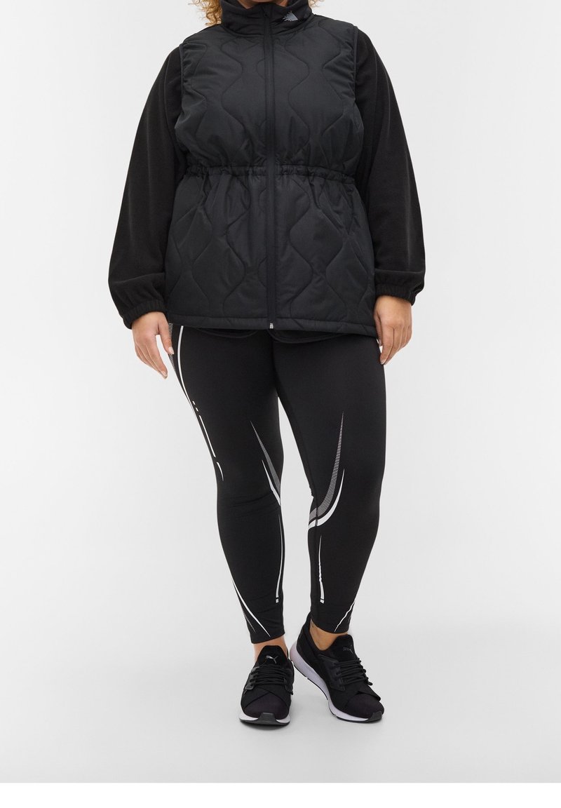Schwarze gesteppte Weste mit hohem Kragen, tailliertem Schnitt und langen schwarzen Ärmeln. Schwarze Leggings mit weißen Akzentstreifen. Schwarze Sportschuhe.