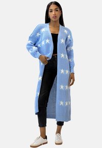 Ljusblå stickad kofta med vita stjärnmönster, öppen framdesign, långa ärmar och ribbade muddar, kombinerad med svarta byxor och sneakers.