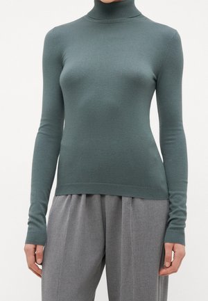 Personne portant un pull à col roulé vert sarcelle à manches longues associé à un pantalon plissé gris ample, sur un fond uni.