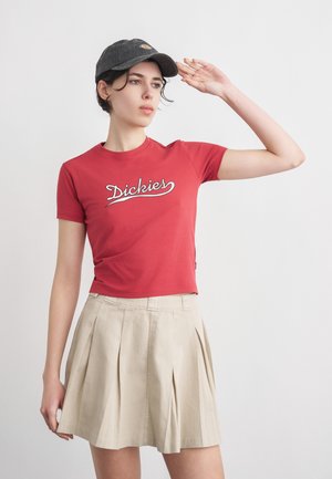 Noor naine, kes kannab punast "Dickies" t-särki, beežist volditud seelikut ja musta mütsi, tõstab ühe käe pea lähedale tavalise tausta ees.