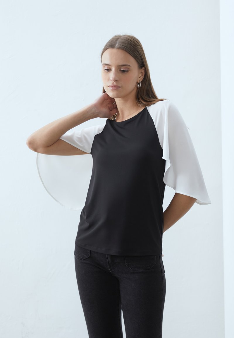 Haut noir avec des manches en chiffon blanc, encolure ronde et coupe décontractée. Tissu lisse avec des manches fluides et un design simple.