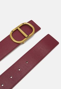 Cintura in pelle rossa con una larga fascia e una grande fibbia circolare in tono oro. Presenta cinque fori per la regolazione della misura.
