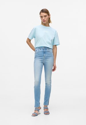 Frau steht und trägt ein hellblaues T-Shirt, hoch taillierte hellblaue Skinny-Jeans mit Knöchelschlitzen und blaue Riemchensandalen vor weißem Hintergrund.