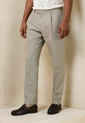 Man wearing light beige tailored trousers and dark brown leather loafers, standing against a beige wall and floor background.