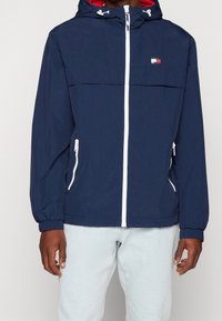 Námořnicky modrá větrovka se bílými zipy, elastickými manžetami a kapucí; uvnitř s červenou podšívkou a malým víceméněbarevným logem na hrudi.