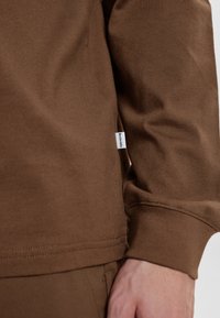 Camicia a maniche lunghe marrone realizzata in tessuto morbido, con una leggera trama e un piccolo etichetta sul lato della cucitura. Dettaglio di cucitura visibile.