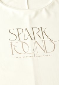 Vaalea beige kangas, jossa on pehmeä rakenne, esittelee painettua tekstiä "SPARK FOUND" tyylitellyllä kultakirjaimilla, ja sen alla lukee "KEEP GROWING | KEEP GOING".