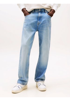 Osoba w jasnoniebieskich prostych spodniach dżinsowych, z rękami w kieszeniach, ubrana w białe trampki, na jednolitym tle.