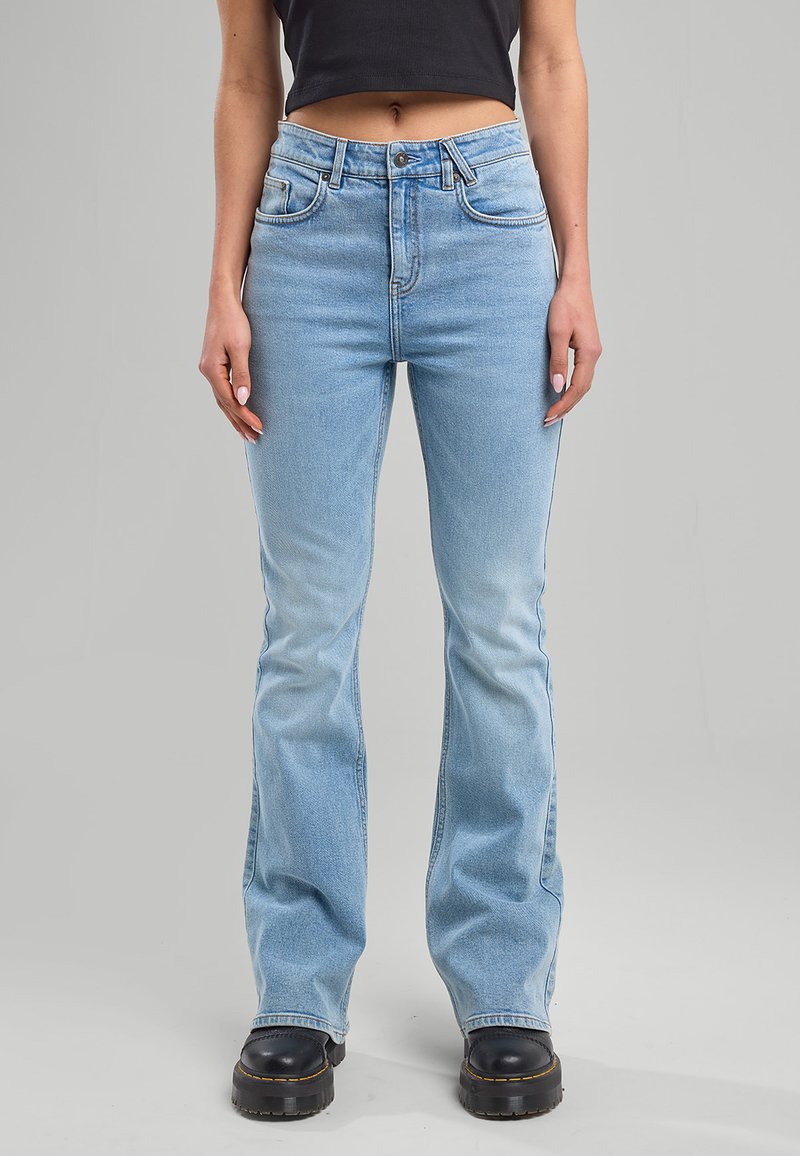 Vrouw draagt lichtblauwe, hooggetailleerde flare-jeans, een zwart cropped topje en zwarte, chunky platformschoenen, staand tegen een effen grijze achtergrond.