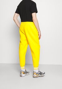 Gelbe Jogginghosen mit elastischem Bund und Bündchen an den Knöcheln, kombiniert mit grau-goldenen Sportschuhen und einem schwarzen T-Shirt.