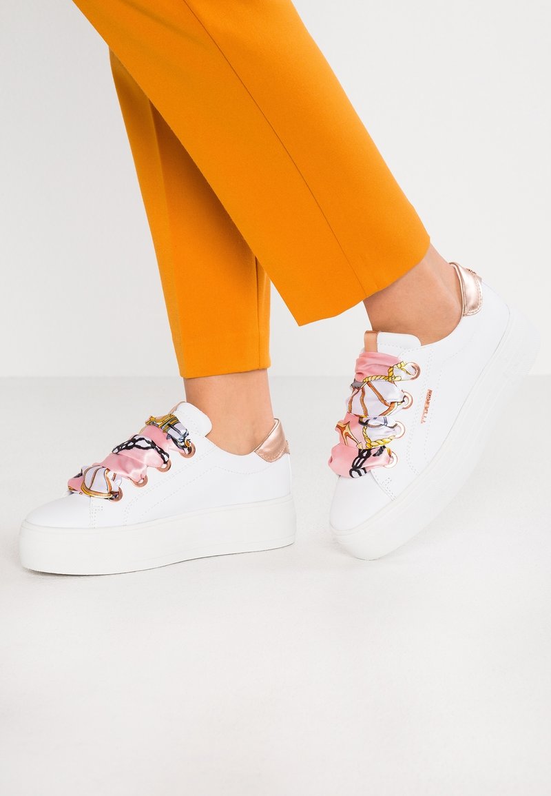 Personne portant des baskets à plateforme blanches avec des lacets en soie rose à motifs et un pantalon orange raccourci sur un fond uni.
