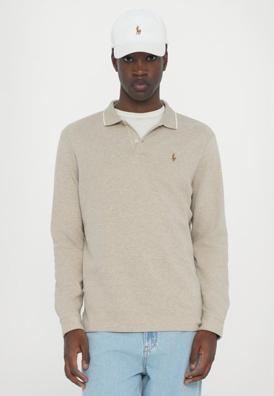 Camisa polo de manga longa bege com colarinho, detalhes brancos, logotipo bordado no peito, combinada com jeans azul claro e um boné branco.