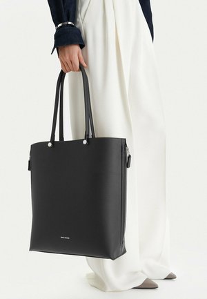 Osoba držící velkou černou tašku za rukojeti, oblečená v bílých širokých kalhotách a tmavém dlouhém rukávu s náramkem.