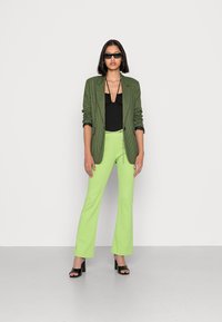 Blazer xadrez verde sobre uma blusa preta, combinado com calças justas e largas de cor verde vivo. Sandálias de salto bloco pretas completam o visual.