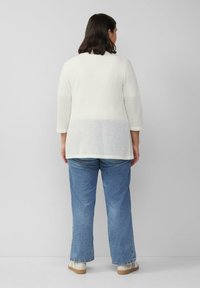 Weißes Strickoberteil mit dreiviertel Ärmeln und strukturiertem Design. Kombiniert mit hellblauen, ausgestellten Jeans und lässigen Slippern.