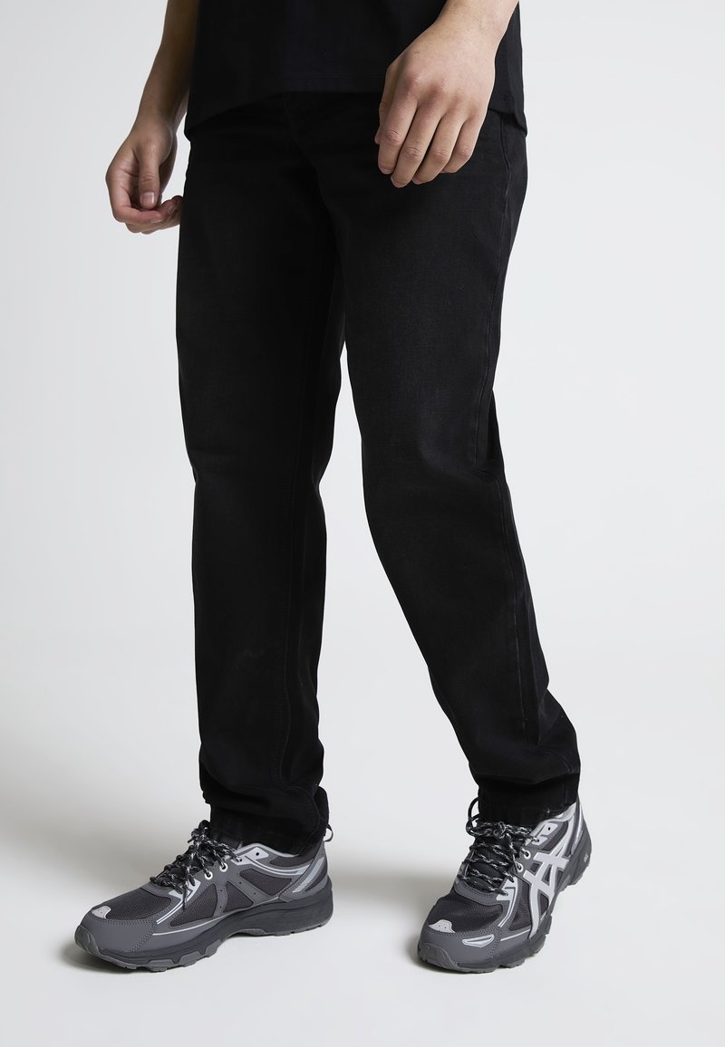 Persona con pantalones negros y zapatos deportivos grises y negros, de pie sobre un fondo claro y liso.