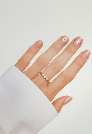 Goldener Ring mit drei klaren Steinen an einer Hand; hellrosa Nagellack mit weißen Blumendesigns; Ärmelsaum eines weißen Langarmhemdes sichtbar.