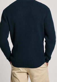 Marineblau gestrickter Pullover mit gerippter Struktur, Rundhalsausschnitt und langen Ärmeln; kombiniert mit hellbeigen Hosen.