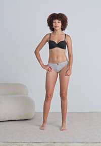 Sutien negru cu sârme și chiloți bikini fără cusături, gri deschis. Țesătură netedă cu detalii minime. Modelul stă pe un covor deschis.