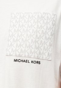 Λευκό βαμβακερό t-shirt με υφασμένη γκρι τσέπη με επαναλαμβανόμενο μοτίβο «MK» και μαύρο κείμενο «MICHAEL KORS» κάτω από την τσέπη.