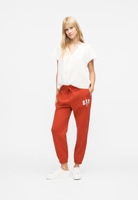 Νεαρή γυναίκα στέκεται φορώντας λευκό πουκάμισο, κόκκινη φόρμα GAP με λογότυπο και λευκά αθλητικά παπούτσια σε λευκό φόντο.