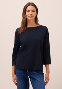 Femme aux longs cheveux bruns portant un haut noir à manches trois-quarts côtelé et un jean bleu, se tenant devant un fond uni.