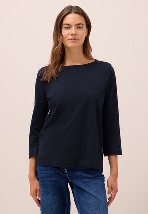Donna con lunghi capelli castani, che indossa una maglietta nera a coste con maniche a tre quarti e jeans blu, in piedi davanti a uno sfondo neutro.