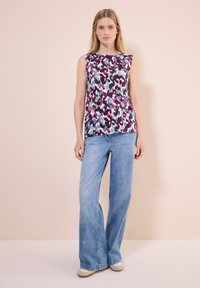 Ärmelloses Oberteil mit einem bunten abstrakten Muster in Pink, Lila und Blau. Kombiniert mit hellblauen, weit geschnittenen Jeans und beigen Schuhen.