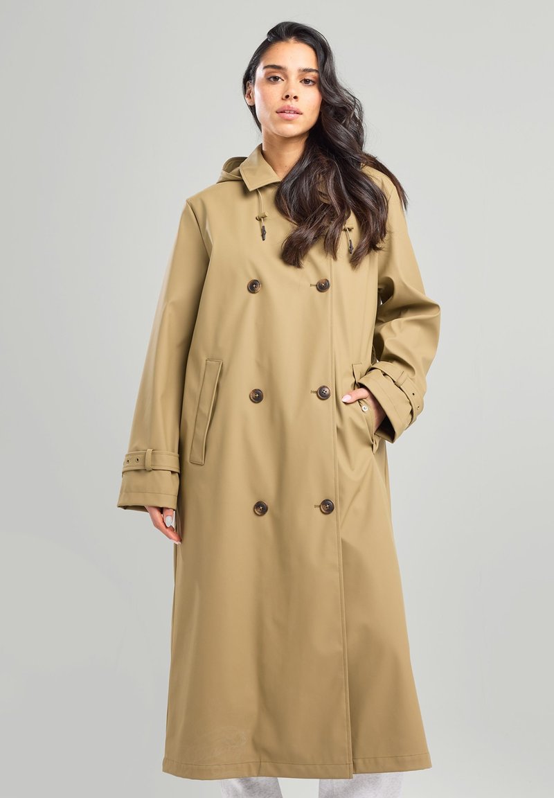 Beige raincoat with double-breasted design, large brown buttons, front pockets, and a hood. Smooth, waterproof material and long sleeves.
