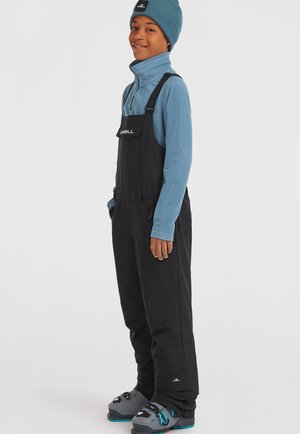 Zwarte skibroeken met schouderbanden en een zak aan de voorkant, gedragen over een blauwe fleece top. Grijze sneeuwlaarzen hebben blauwe accenten en gespen.