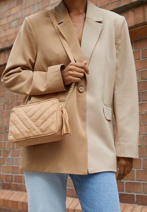 Bolso de mano acolchado en tono marrón con un logo dorado y un fleco, llevado bajo un blazer oversized beige y marrón claro con un solo botón.