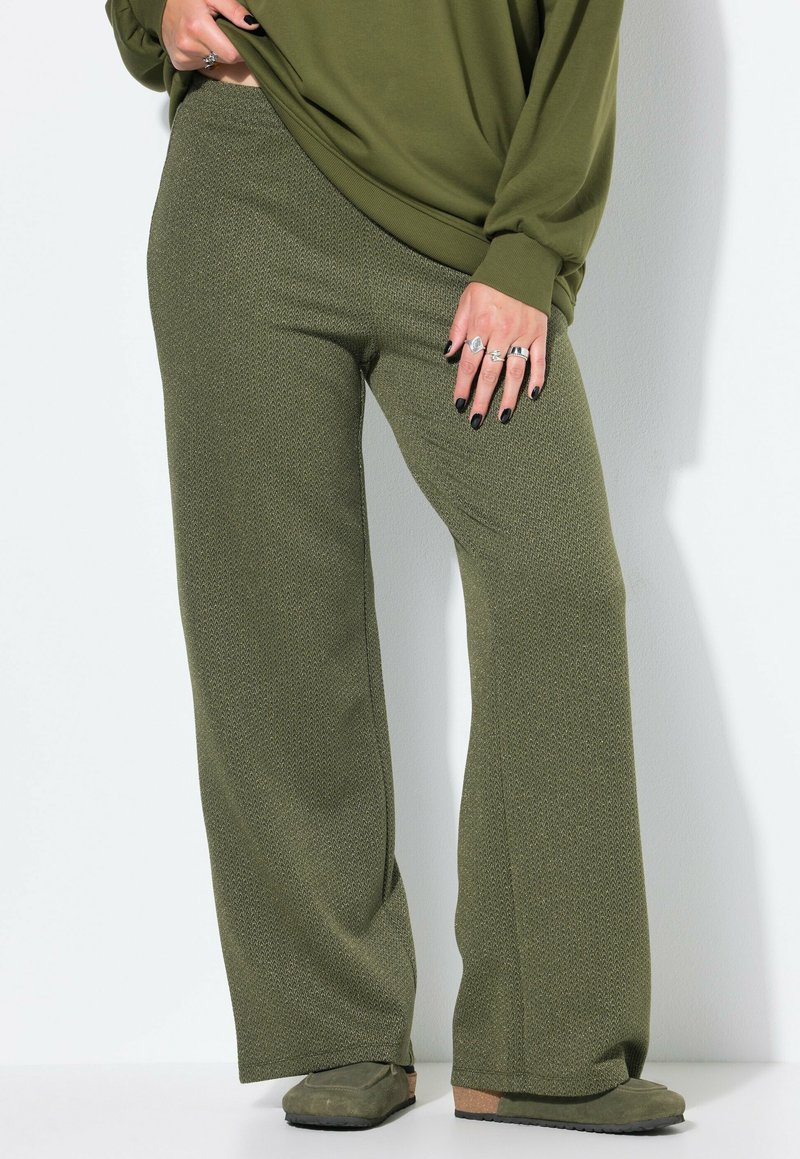 Vrouw in olijfgroene gebreide brede broek, bijpassende losse top, meerdere ringen en groene instappers die tegen een witte muur staat.