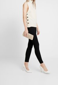 Krämfärgad ärmlös topp med sidoknappar, kombinerad med svarta skinny jeans och vita högklackade skor. Håller en guldfärgad clutch med glitter.