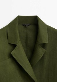 Blazer in lino verde oliva con colletto a revers, taschino sul petto e dettagli di cucitura visibili lungo i bordi.