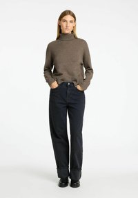 Brauner Strickpullover mit Stehkragen, kombiniert mit dunklen Jeans. Die Jeans haben einen lockeren Schnitt und umgekrempelte Säume. Schwarze Stiefeletten runden das Outfit ab.