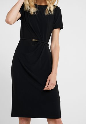 Vrouw die een korte zwarte jurk draagt met een smalle riem in de taille, staand tegen een lichte effen achtergrond.