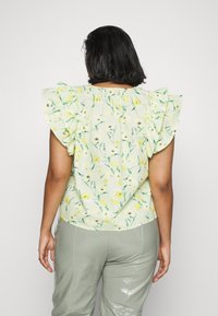 Blomstermønstret bluse i lys grøn med flæser på ærmerne, med gule og sorte detaljer. Glat stof med en afslappet pasform.