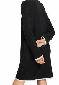 Robe noire tricotée avec poignets et ourlet côtelés blancs. Présente une coupe décontractée et des manches longues, vue de profil.