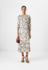 Beige Midikleid mit floralen Druck in Schwarz mit botanischen Formen, ausgestattet mit einem hohen Kragen und Puffärmeln. Kombiniert mit einer kleinen roten Tasche und schwarzen High Heels.