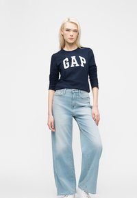 Une femme blonde porte un sweat-shirt marine GAP, un jean large bleu clair et des baskets blanches, se tenant devant un fond blanc uni.