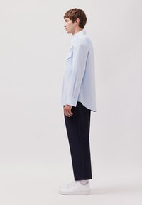 Camicia a maniche lunghe azzurro chiaro con colletto a button-down, taschino sul petto, abbinata a pantaloni scuri e sneakers bianche. Texture liscia, vestibilità moderna.