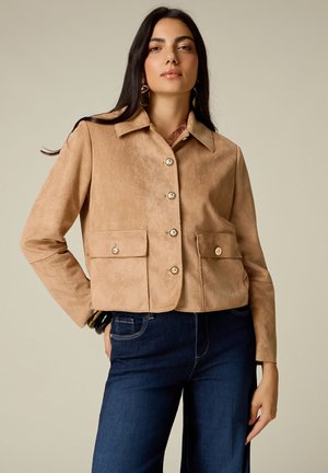 Femme portant une veste en suède beige à boutons avec poches à rabat et un jean bleu foncé, debout avec une main dans la poche devant un fond uni.