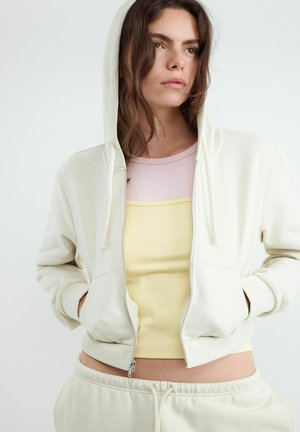 Jeune femme aux cheveux bruns ondulés portant un sweat à capuche crème zippé, un haut jaune pastel et un pantalon de survêtement crème sur fond uni.